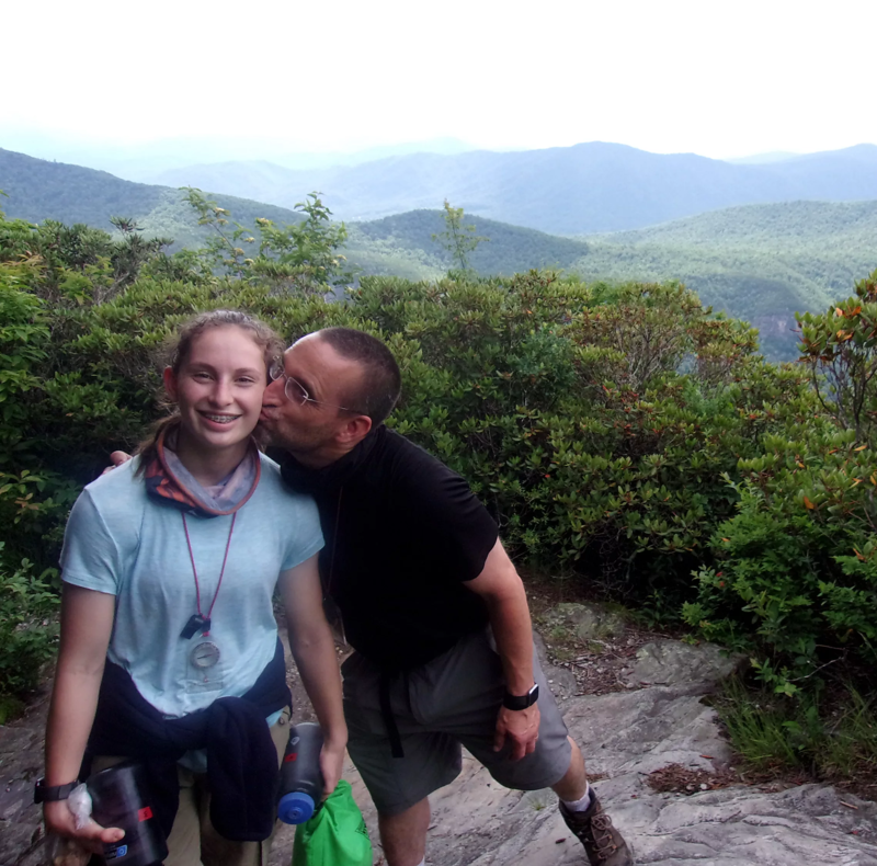 A man is kissing a young woman on the cheek while standing on a mountain trail. The woman is wearing a light blue shirt, and the man is wearing a black shirt. They are surrounded by lush greenery, and in the background, there are rolling hills and mountains. The sky is overcast, creating a soft, diffused light. The photo captures a candid moment of affection amidst a scenic outdoor setting.